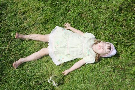 光着脚的小女孩躺在草甸在夏日的阳光下的绿草照片