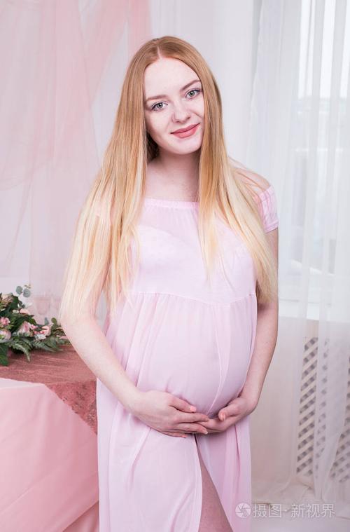 孕妇抚摸她的腹部和微笑.妇女在怀孕期间粉红色礼服在粉红色和白色的